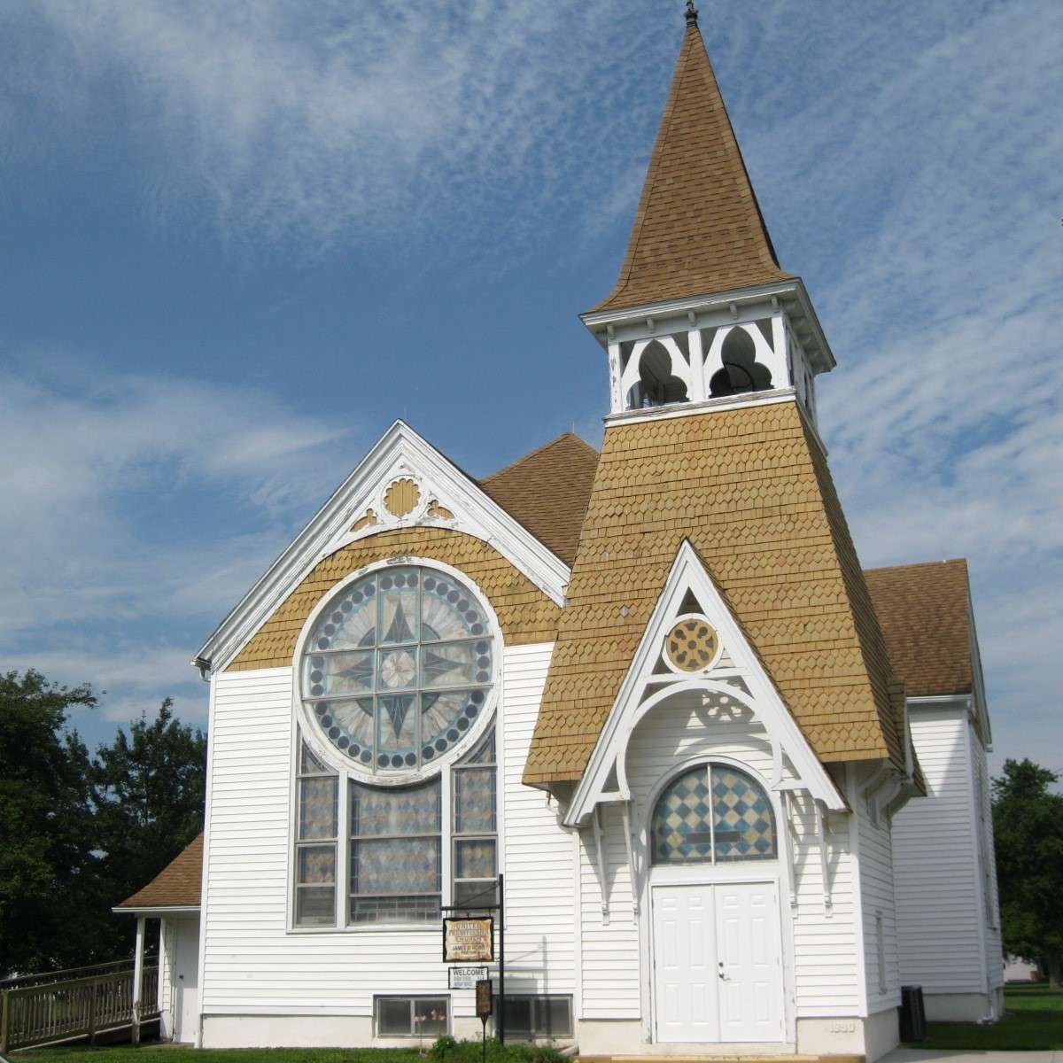 Two story wooden church building with white wooden siding, brown shingled roof, stained glass windows, steeple tower with bell