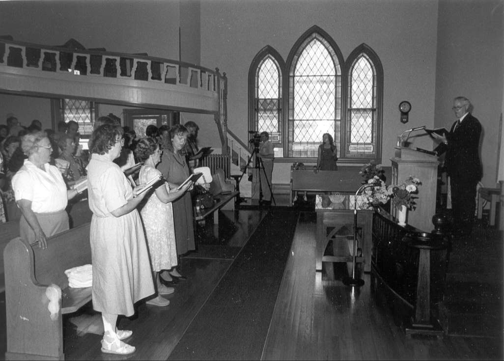 black and white image of older gentleman preaching from book at pulpit in small church with stained glass windows