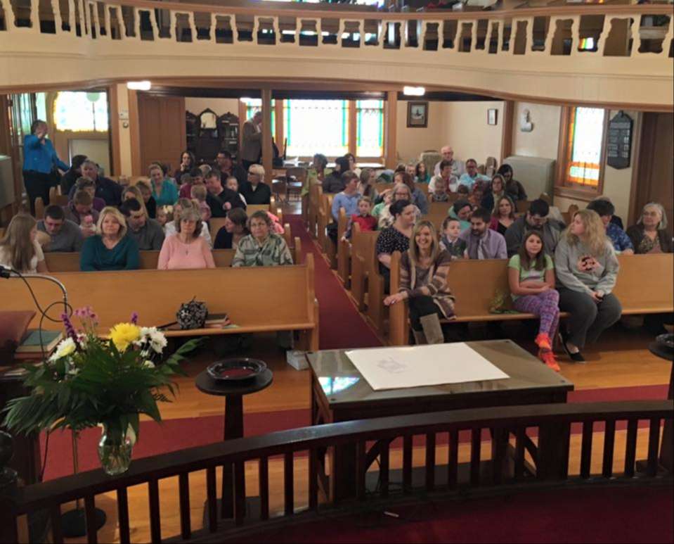 church sanctuary pews full of people of all ages and genders, curved balcony above their heads, communion table and curved pulpit in foreground